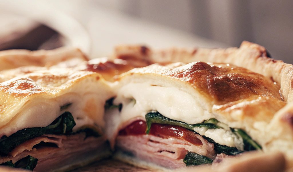 Golden baked Pizza Rustica with a crisp crust, cut open to reveal layers of spinach, mozzarella, fontina cheese, cherry tomatoes, and thin slices of turkey ham, served in a rustic pie dish.
